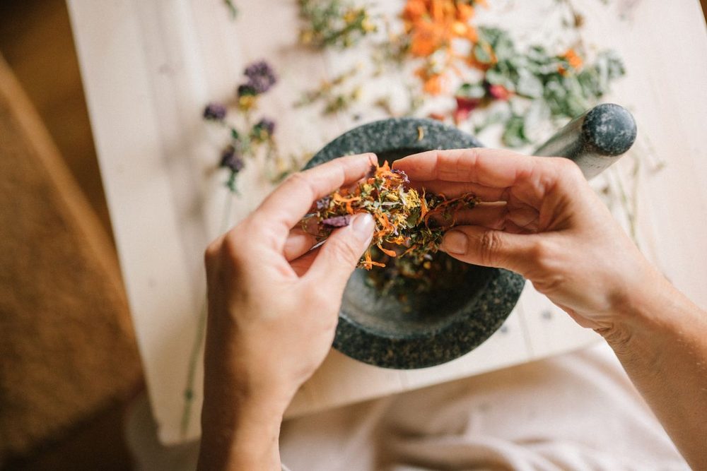 Hands blending dried herbs and flowers over a mortar and pestle — daily Ayurvedic practices for home